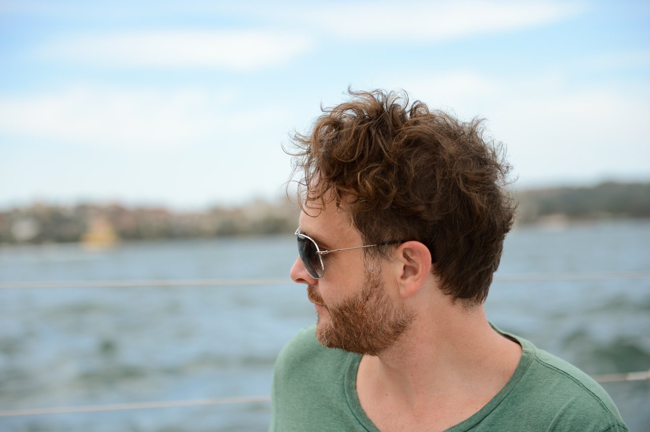 A man with curly brown hair, a beard, and sunglasses looking to the side.