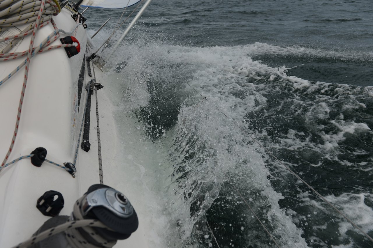 A close-up view of a sailboat cutting through choppy water, with foamy waves splashing against the white hull, ropes, and rigging under an overcast sky.