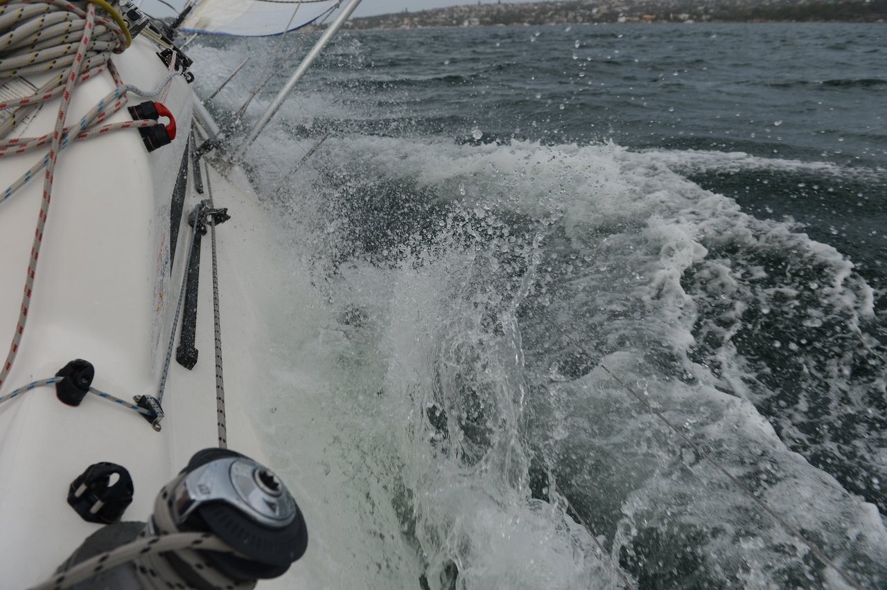 Water splashes against the side of a sailing boat, which is tilted at an angle with ropes, pulleys, and rigging visible, as choppy waves and a distant shoreline appear in the background.