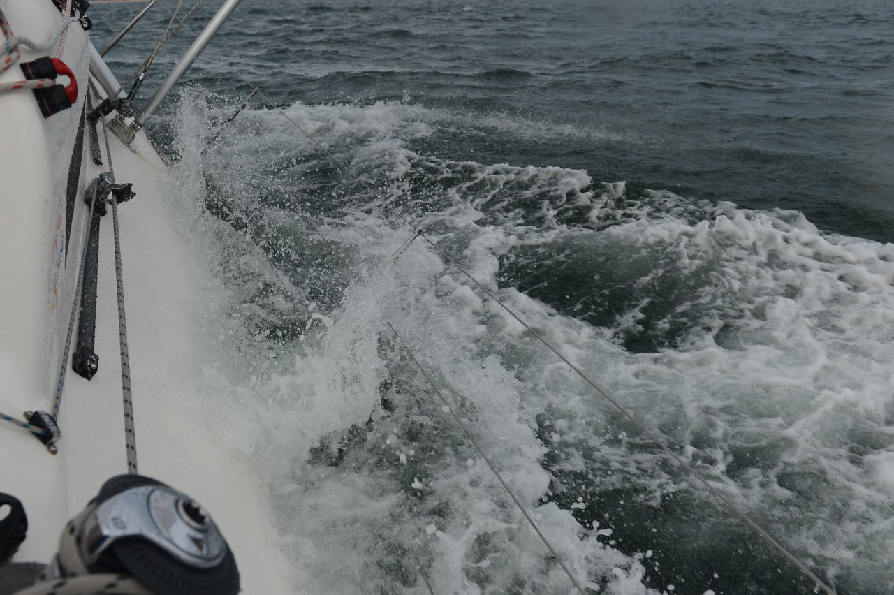 Water splashes against the side of a sailing boat, with ropes, metal rigging, and a winch visible in the foreground, as the boat moves through choppy waves.