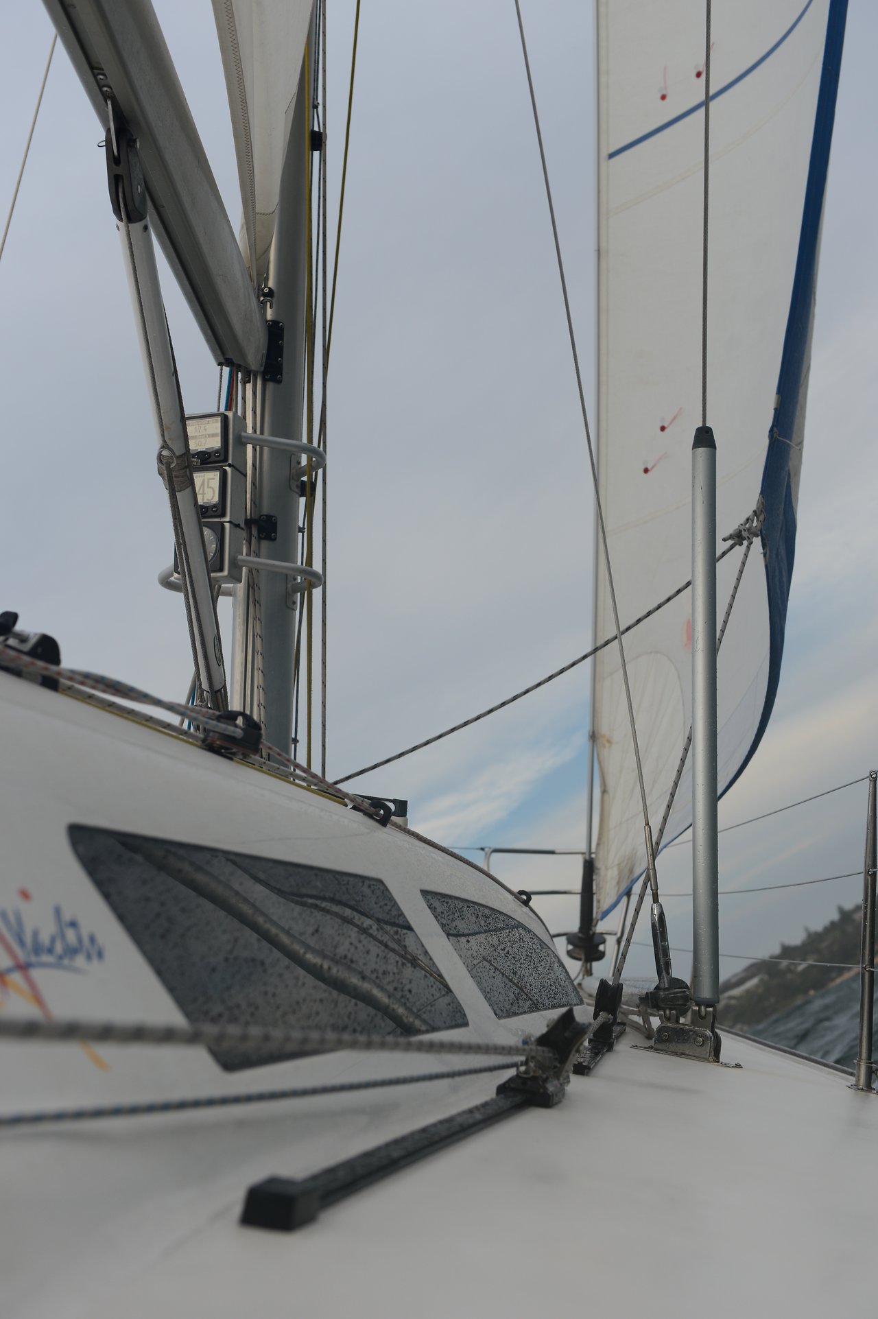 A close-up view of a sailboat's deck shows wet cabin windows, taut ropes, and tall white sails against a cloudy sky, with a distant shoreline visible on the right.