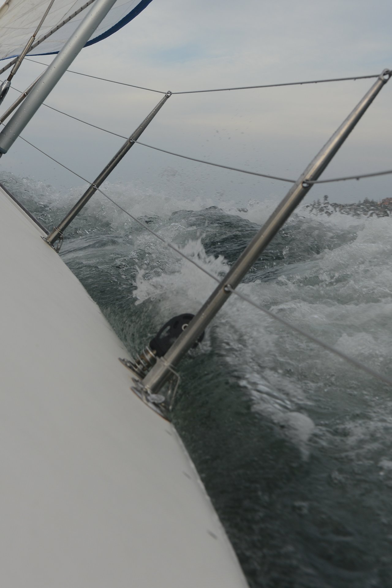 A close-up view from the side of a sailboat shows metal railing wires and hardware as waves crash against the tilted hull under a cloudy sky.