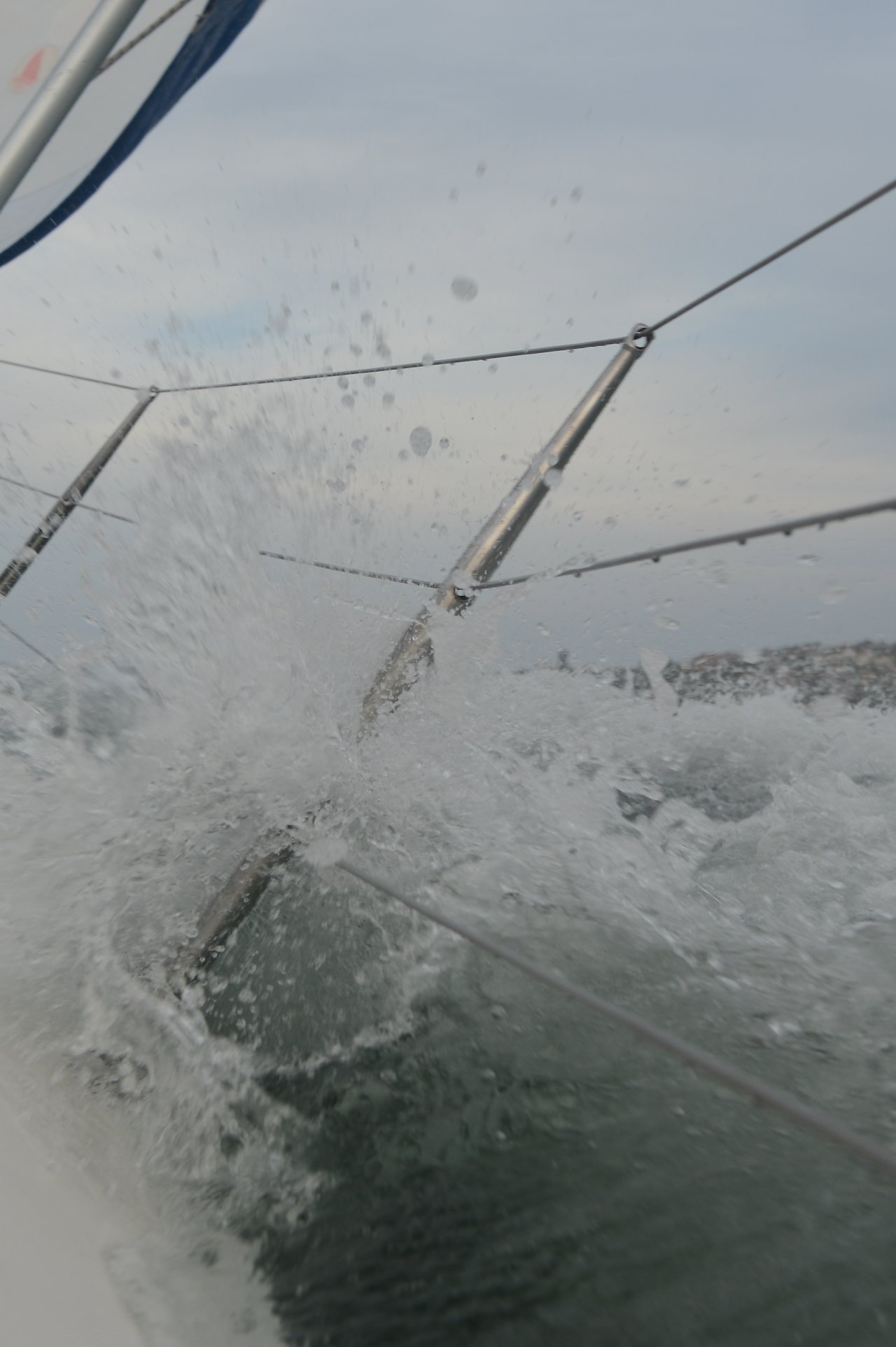 Water splashes against the metal railing of a sailboat, with taut ropes stretching diagonally.