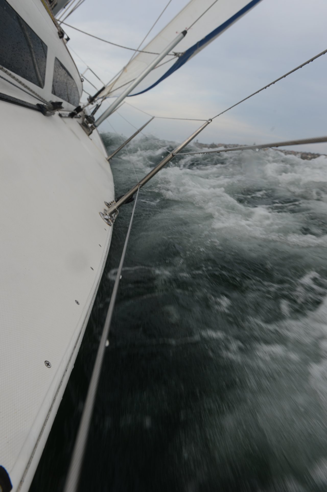 A sailboat leans sharply to the left as it moves through rough, choppy waters, with white sails stretched above and metal rigging lines cutting across the frame.