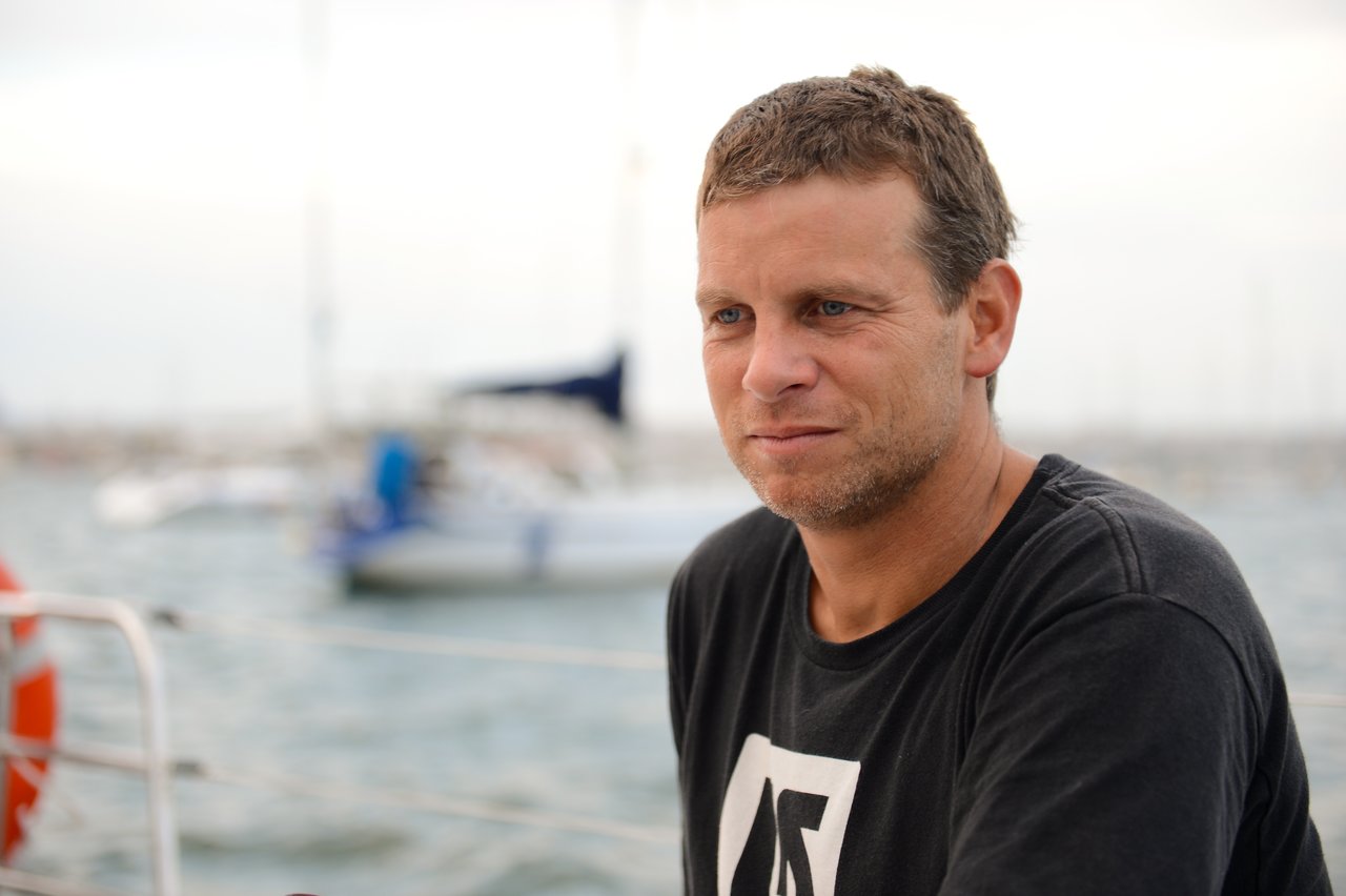 A man with short, light brown hair and stubble wears a black T-shirt with a white logo, sitting on a boat with a blurred marina and sailboats in the background.