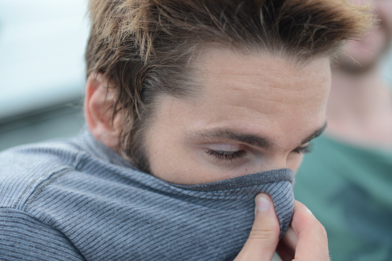 A man pulls up the collar of his gray shirt to cover his nose.