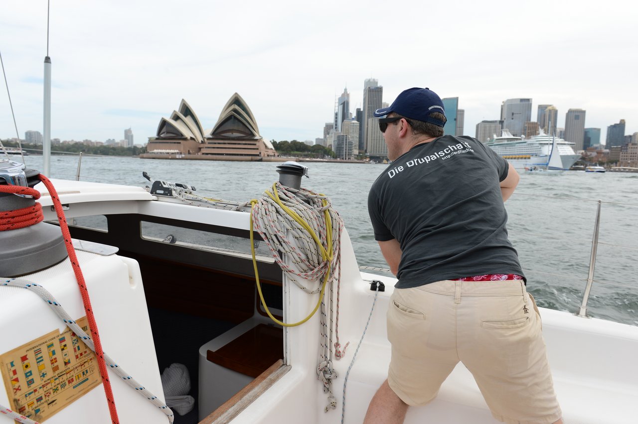 A man wearing a dark t-shirt and beige shorts is adjusting a rope on a sailboat, with the Sydney Opera House, city skyline, and a cruise ship in the background.