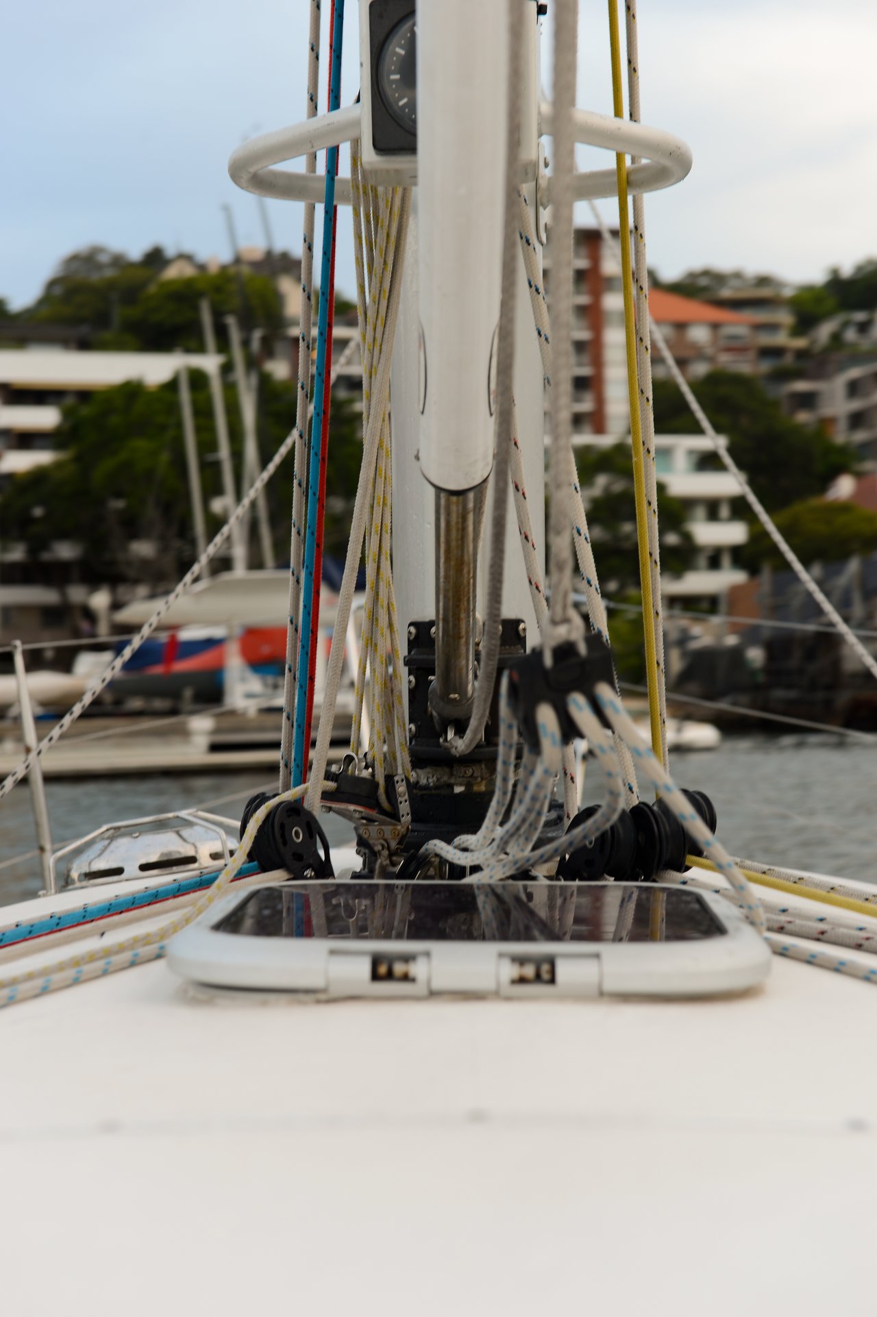 Close-up view from the deck of a sailboat, showing the mast, rigging, and ropes, with a marina, and docked boats.