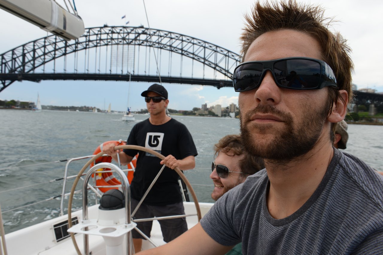 Three men are on a sailboat near a large steel bridge, with one steering.