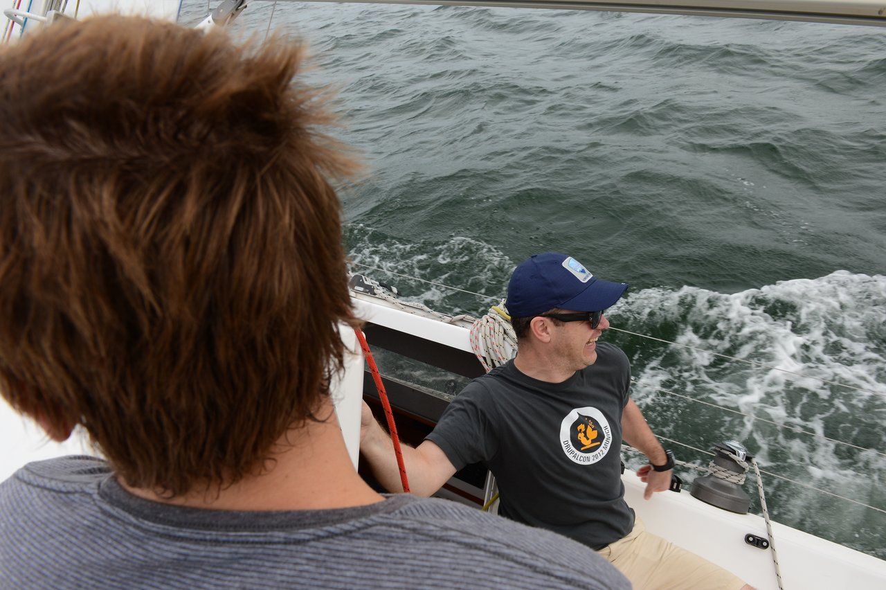 Two people are on a sailboat, with one in the foreground facing away and another wearing a cap and sunglasses, smiling while holding a rope near the boat's edge as water splashes behind.