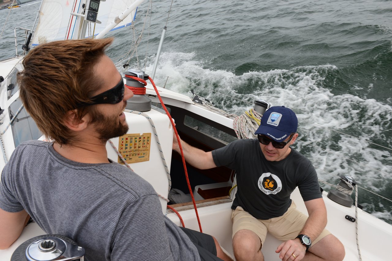Two men on a sailboat hold on as the boat tilts steeply.
