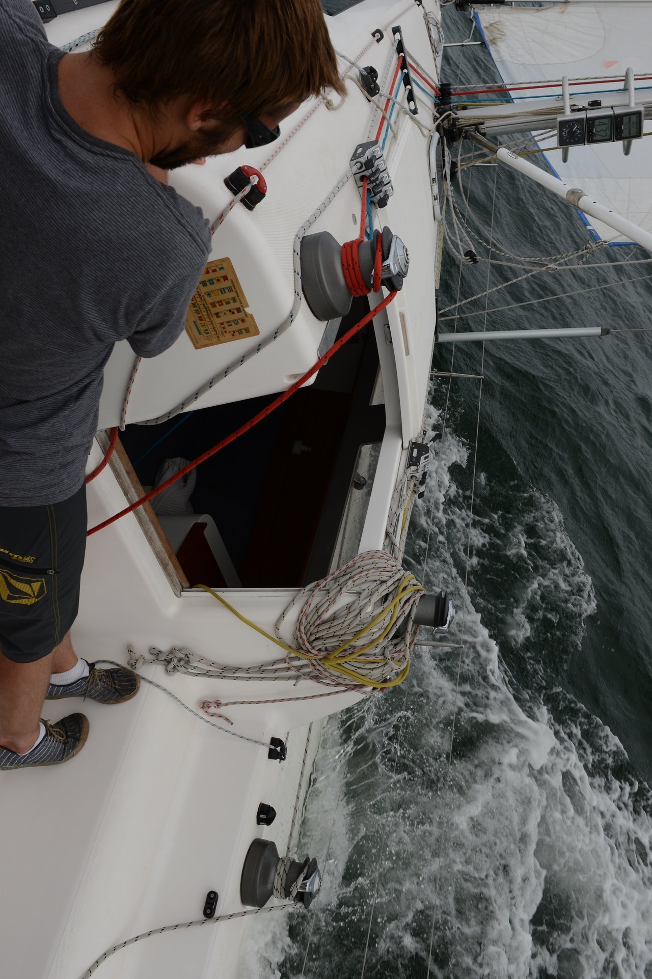 A man wearing a gray shirt and sunglasses leans over the tilted deck of a sailing boat as waves crash against the hull.