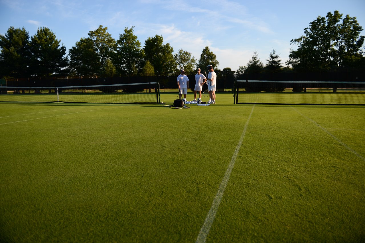 Four people stand near the net on a grass tennis court, talking and preparing for a match.