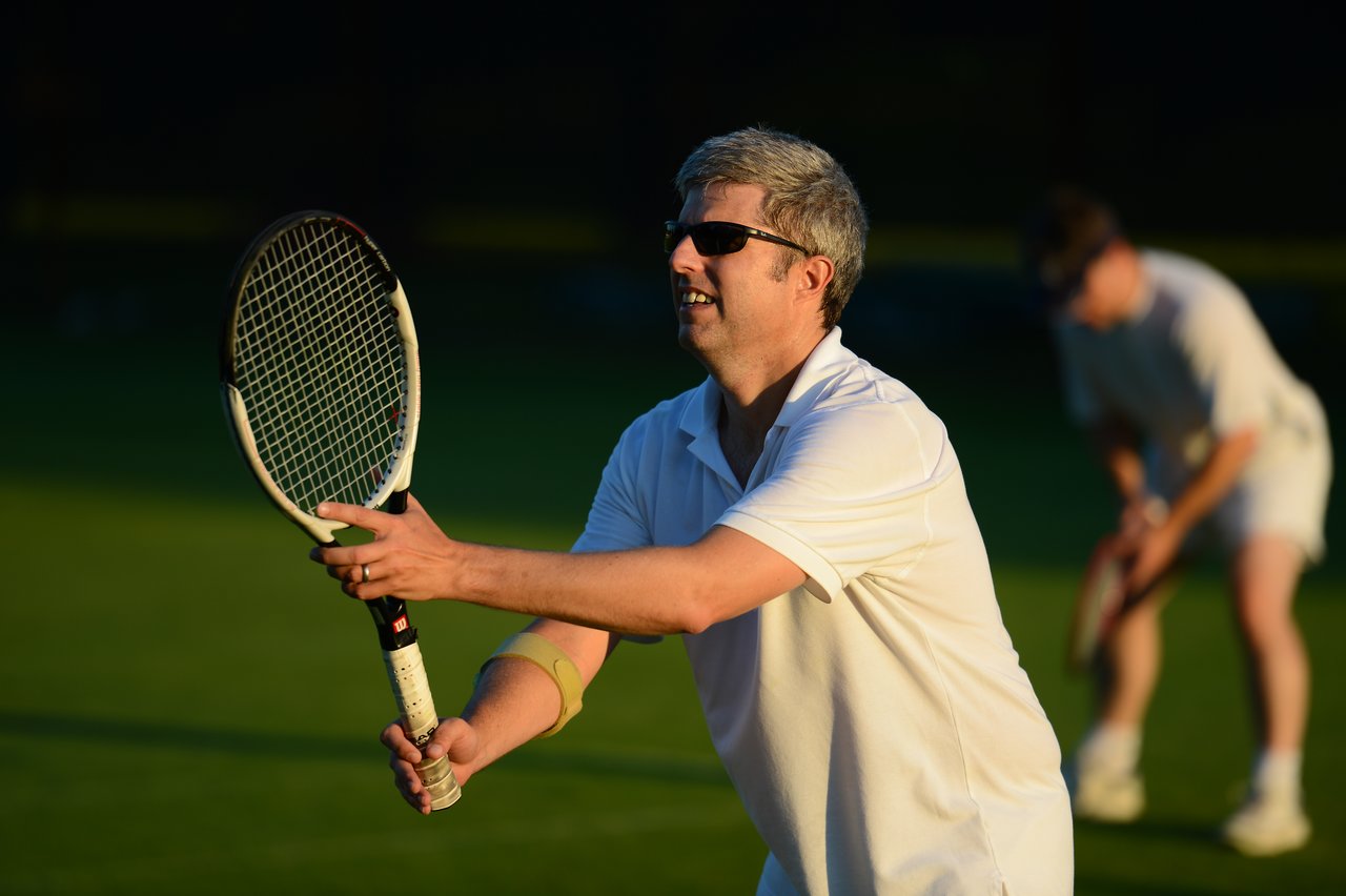 A man in a white outfit and sunglasses prepares to hit a tennis ball with his racket.