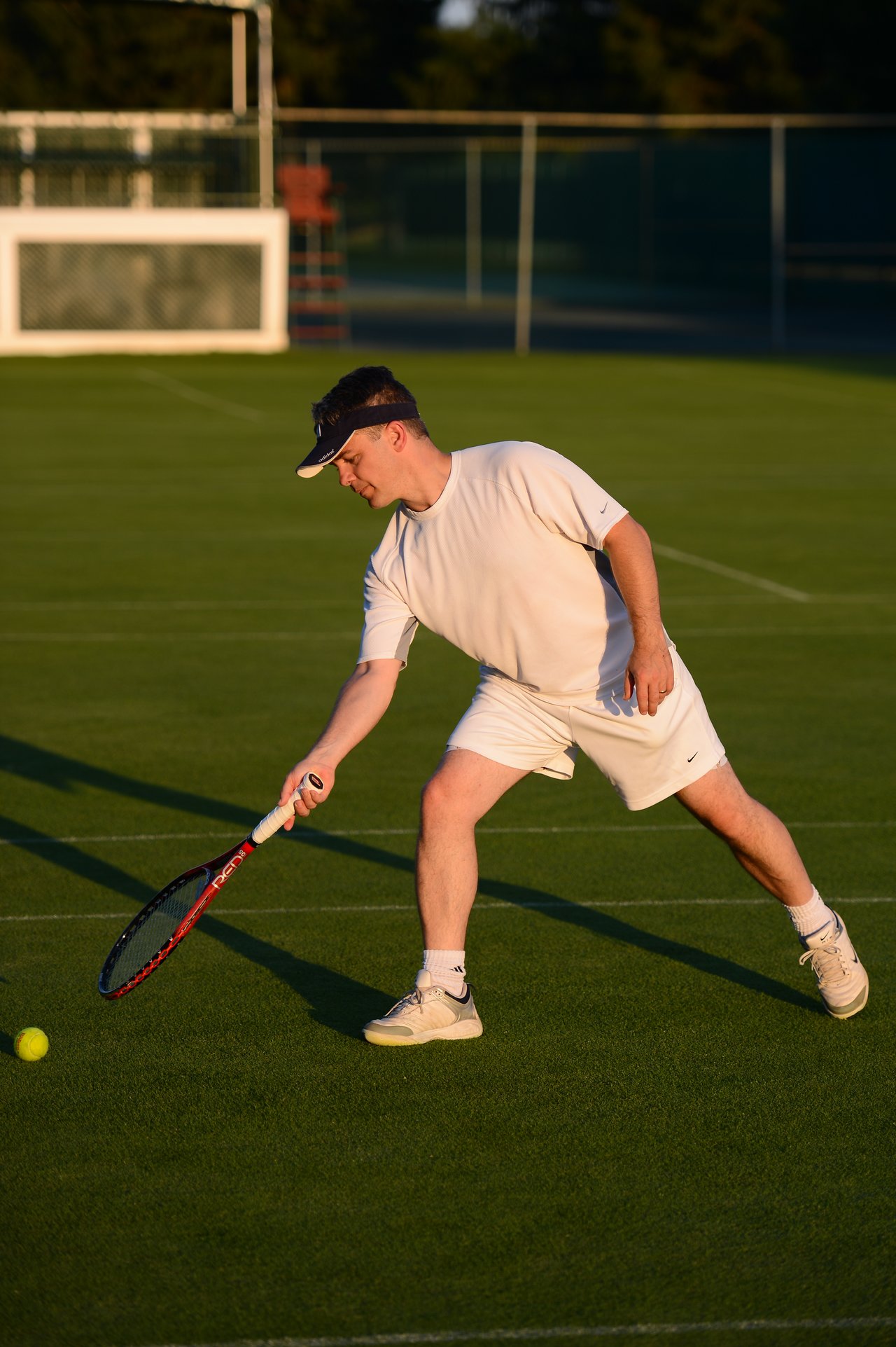 A tennis player in white reaches to hit a low ball with a racket on a grass court.