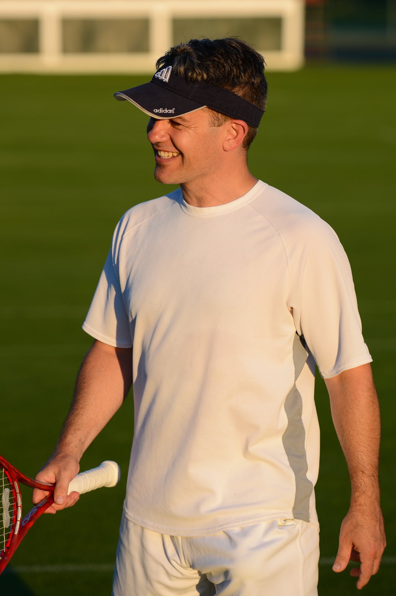 A man in a white tennis outfit holds a racket and smiles while standing on a grass court.