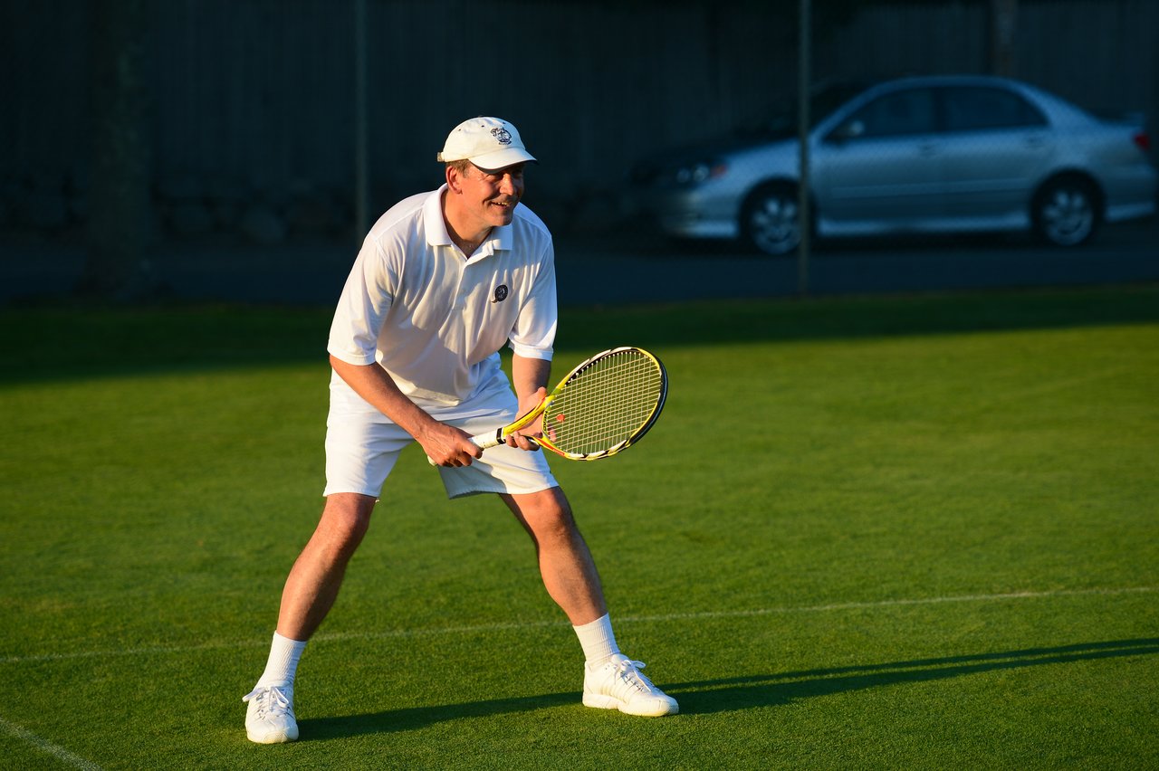 A tennis player in white attire holds a racket and prepares to receive a shot on a grass court.