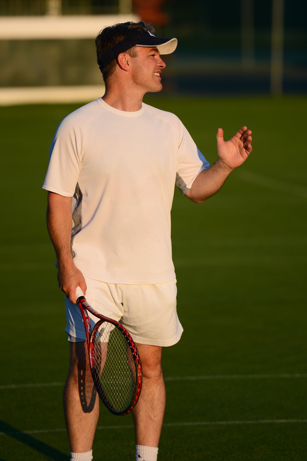 A man in a white outfit holds a tennis racket and gestures while standing on a grass court.