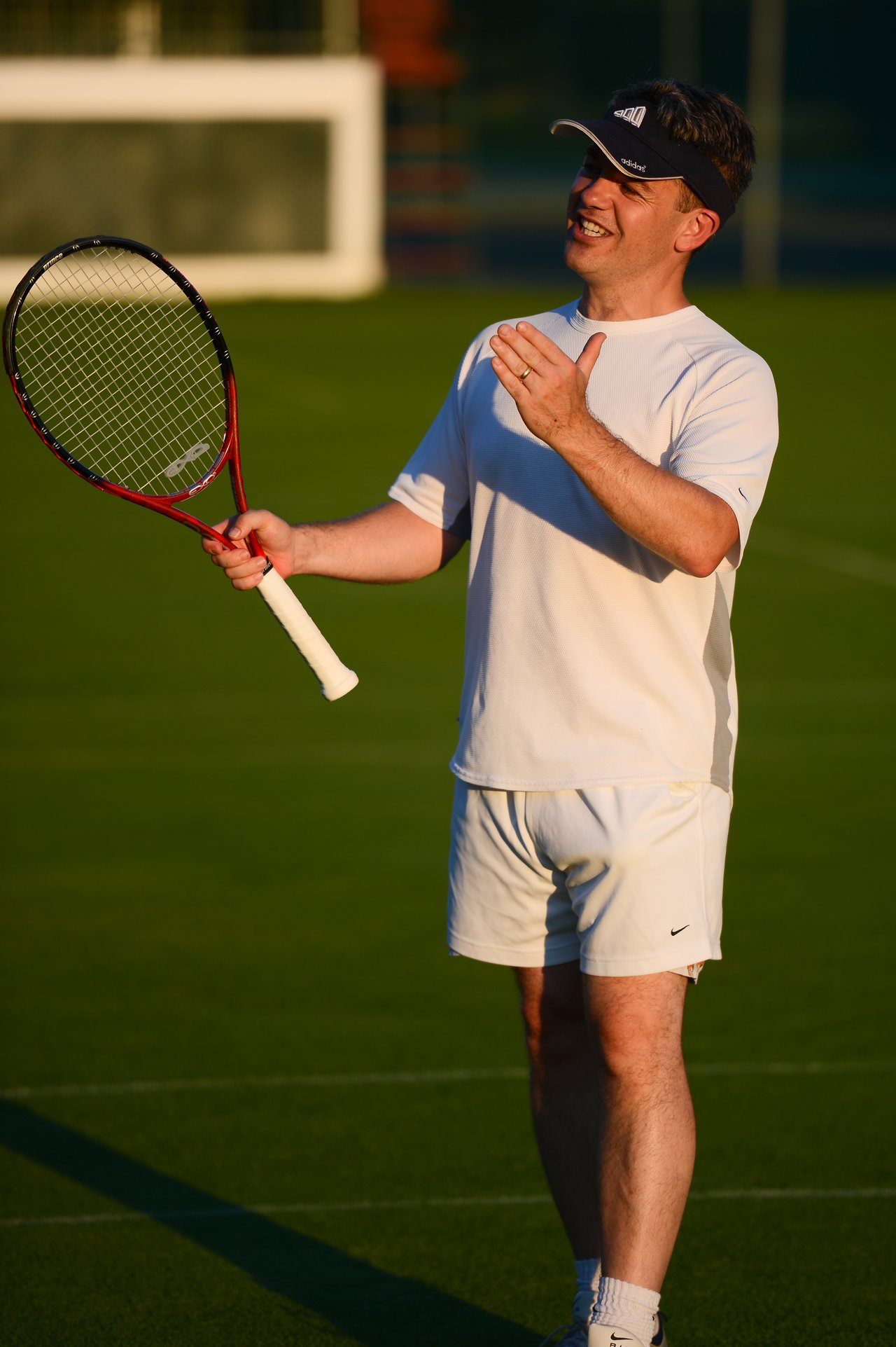 A man in white tennis attire holds a racket and gestures while standing on a grass court.