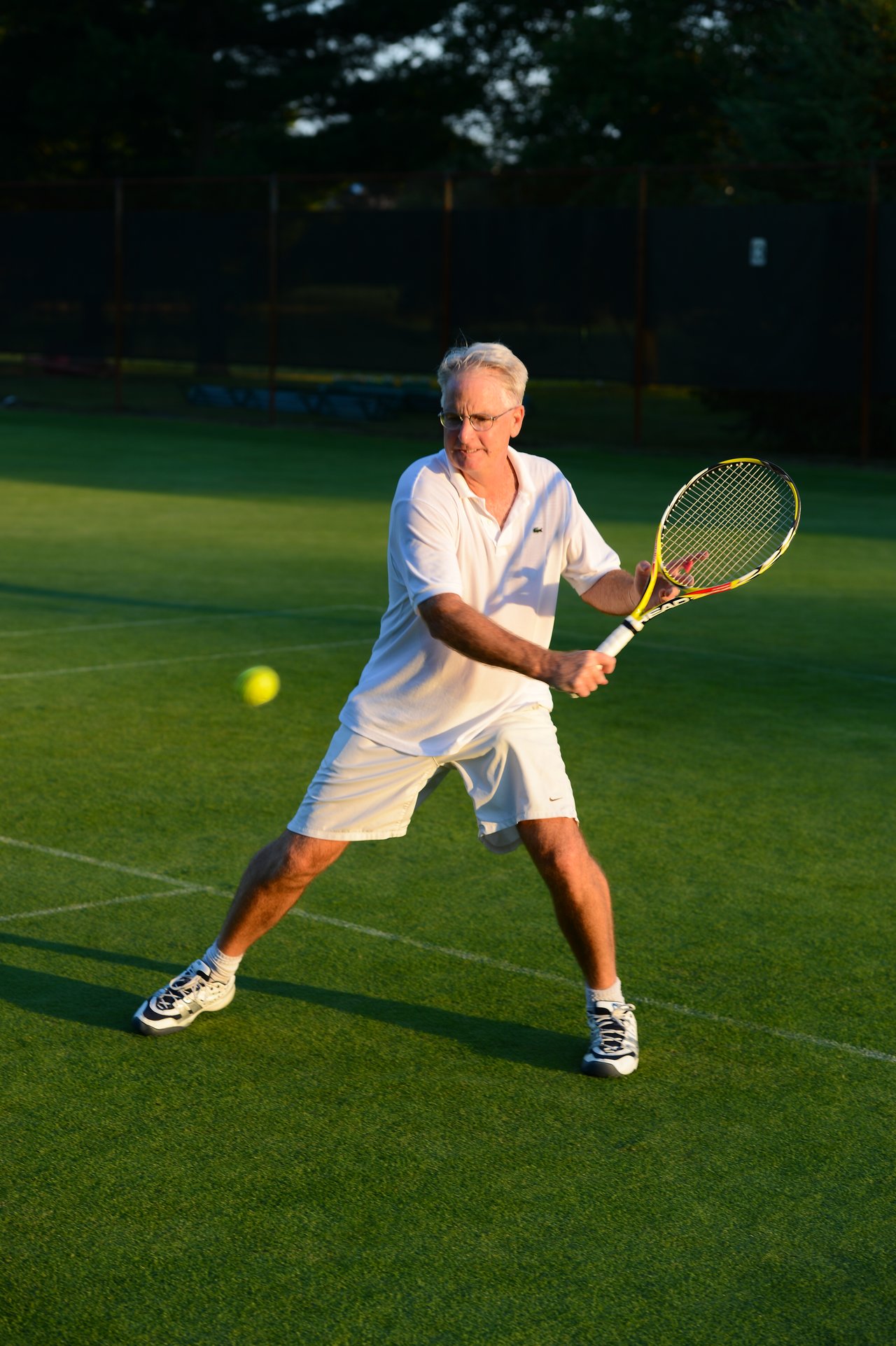 A man in white sportswear prepares to hit a tennis ball with a backhand stroke on a grass court.