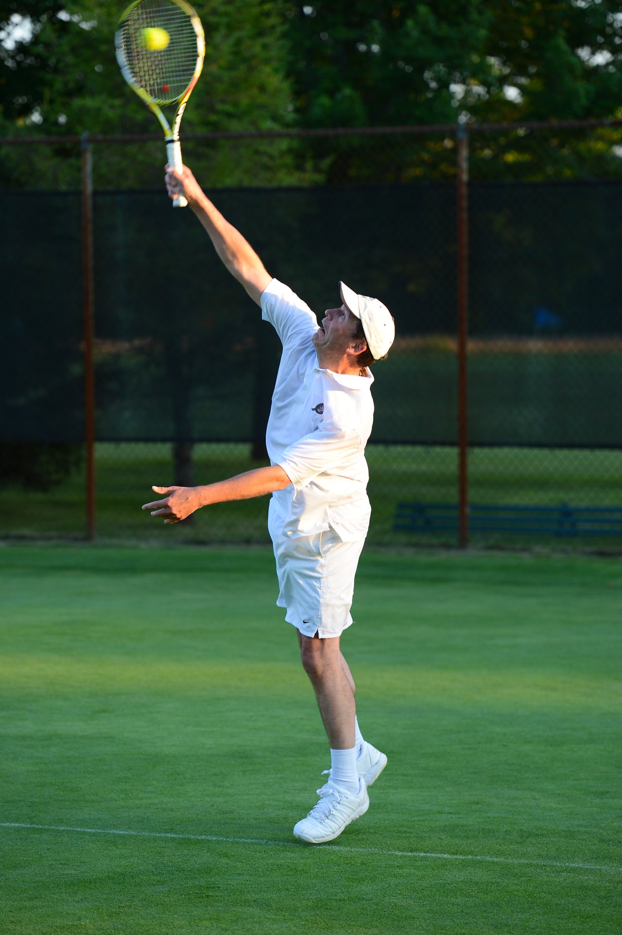 A tennis player in white clothing serves the ball on a grass court, holding the racket overhead.