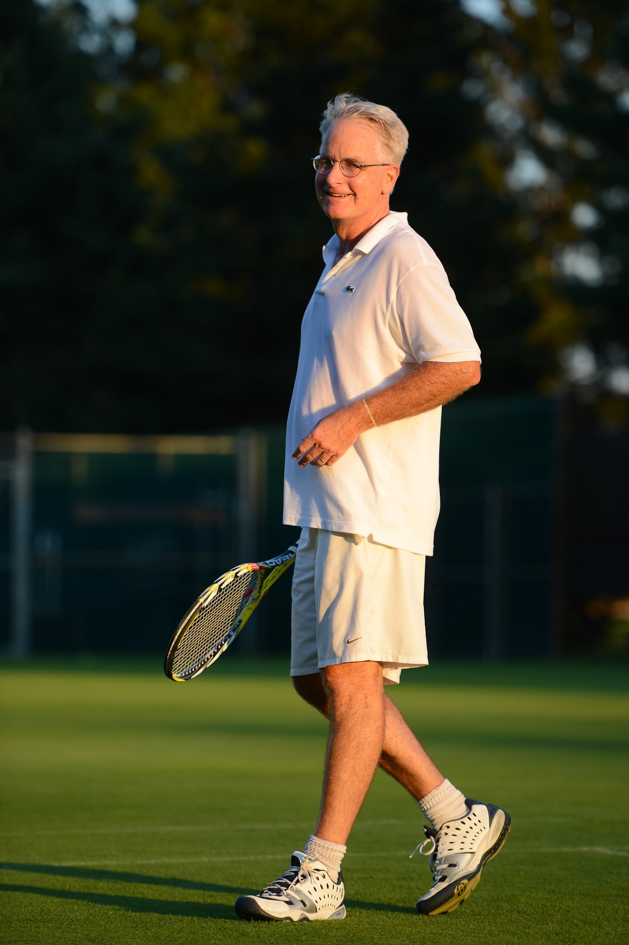 A man in white tennis attire walks on a grass court, holding a tennis racket and smiling.
