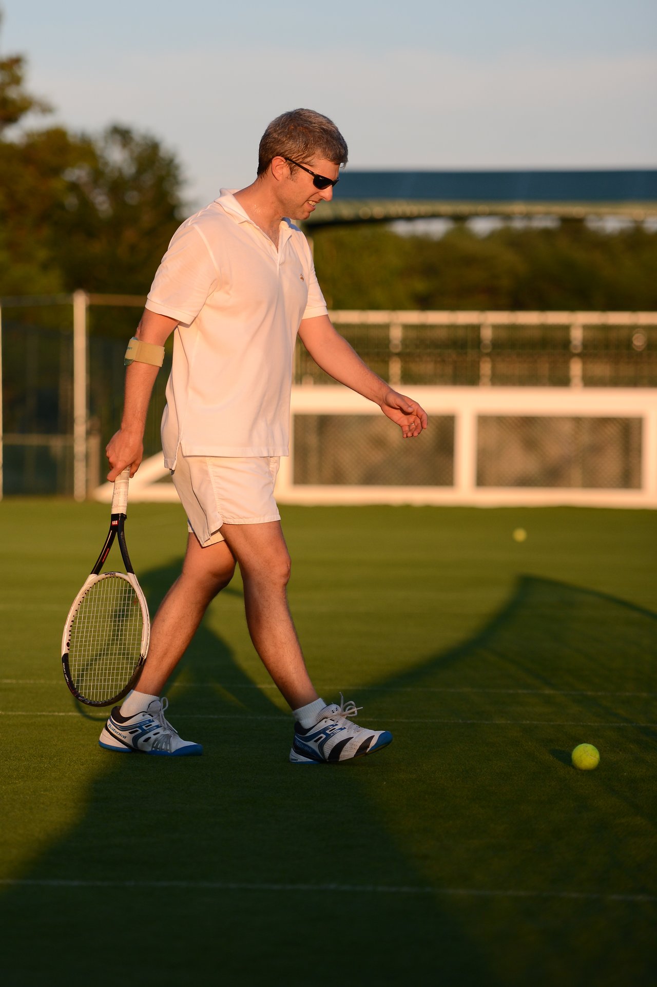 A man in white tennis attire walks on a grass court, holding a racket and approaching a tennis ball.