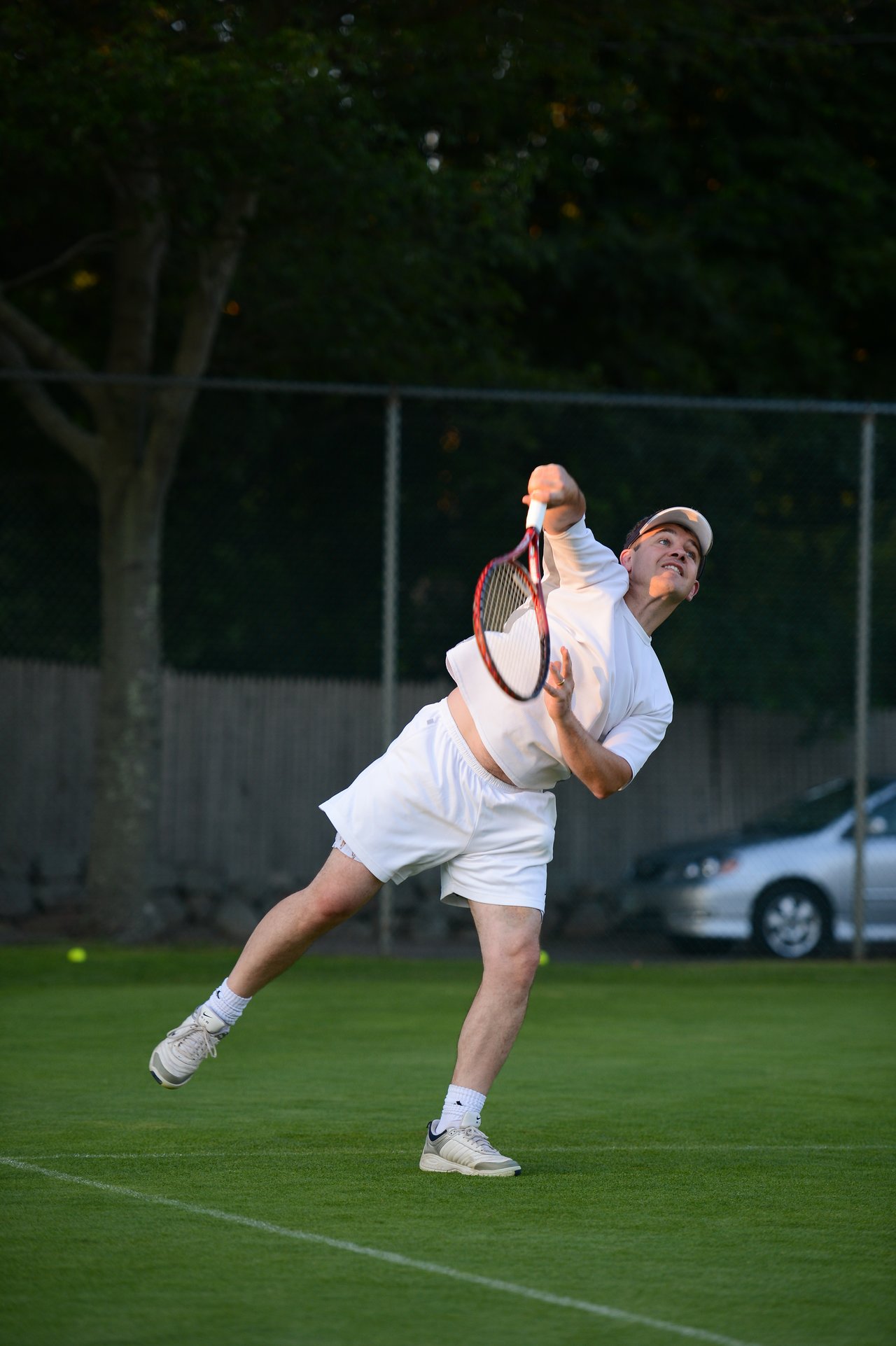 A tennis player in white serves the ball on a grass court, holding a racket with an extended arm.