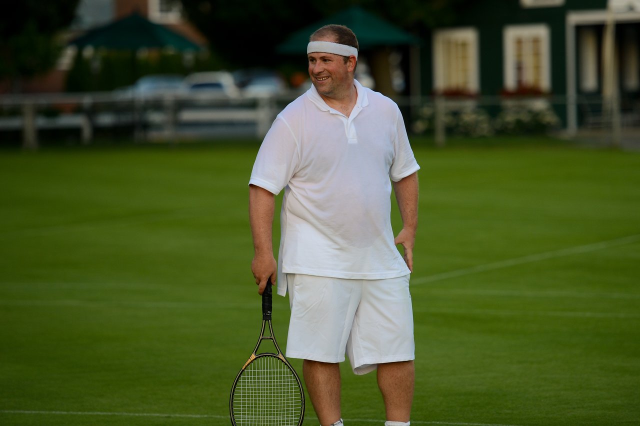 A man in white tennis attire stands on a grass court, holding a racket and smiling.