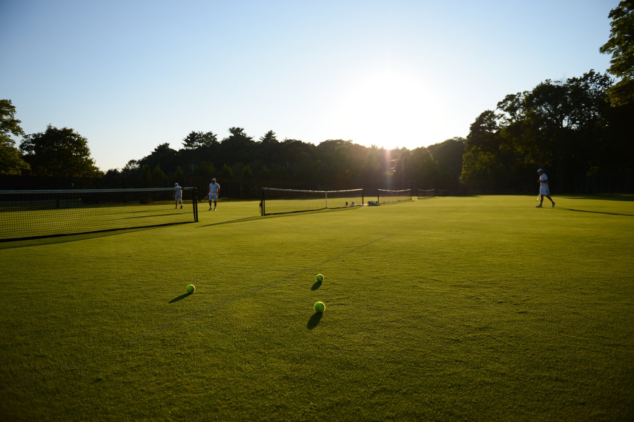 Tennis players in white outfits walk on a grass court with tennis balls scattered in the foreground.