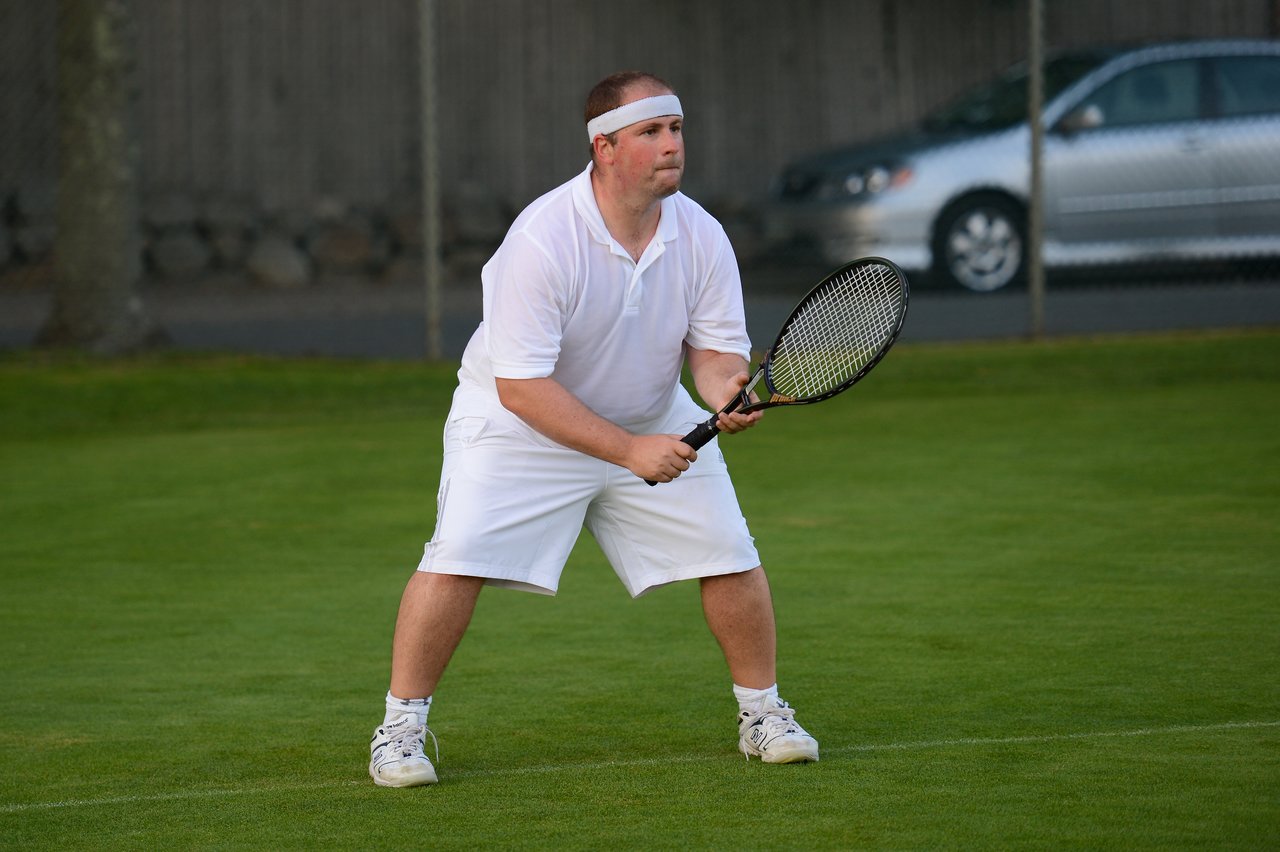A tennis player in white attire holds a racket and prepares to receive a shot on a grass court.