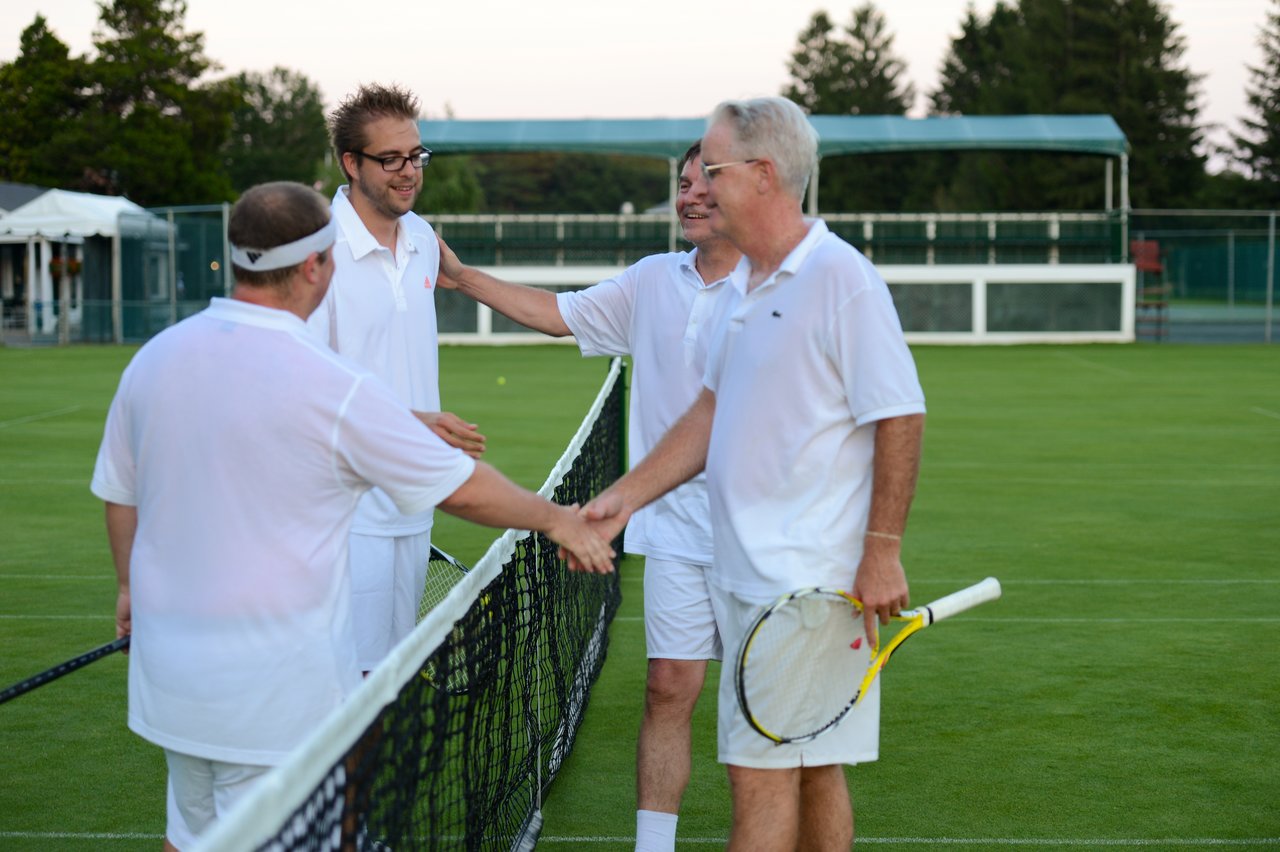 Four men in white tennis outfits shake hands at the net after a match on a grass court.
