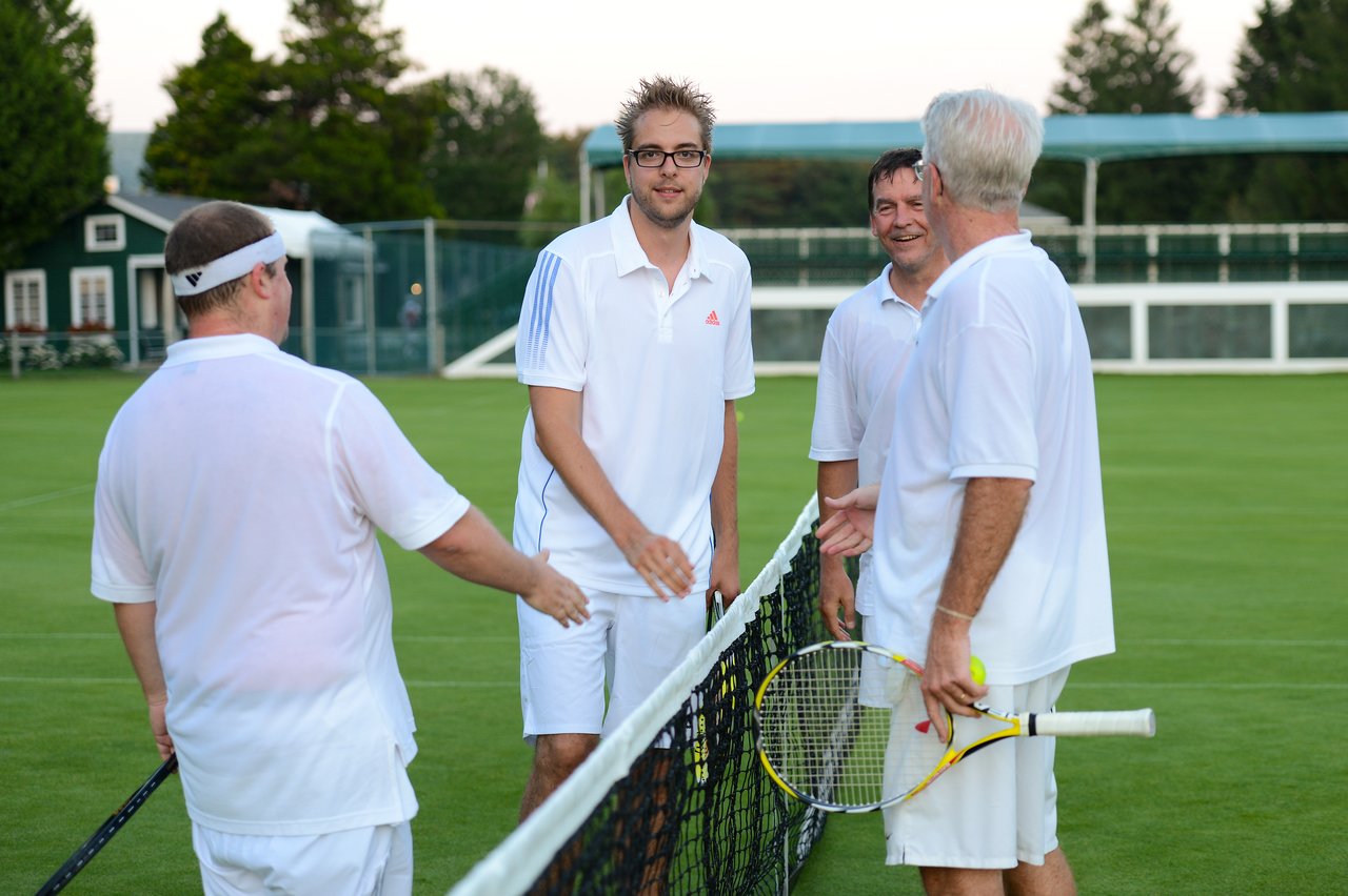 Four men in white tennis outfits stand at the net, shaking hands and talking after a match.