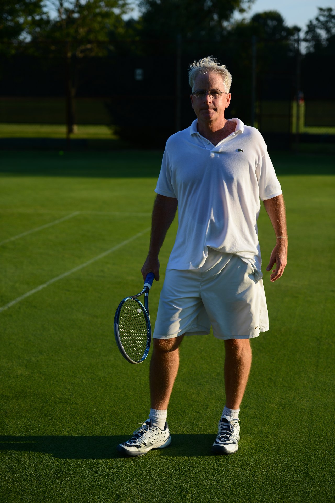 A man in a white tennis outfit stands on a grass court, holding a tennis racket in one hand.