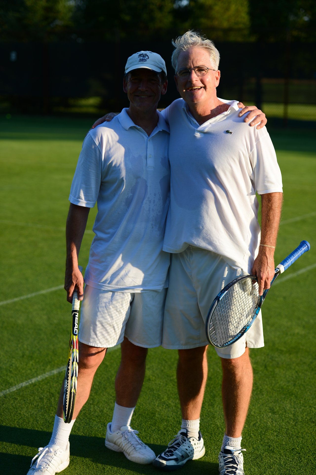 Two men in white tennis outfits stand on a grass court, smiling and holding rackets after a match.