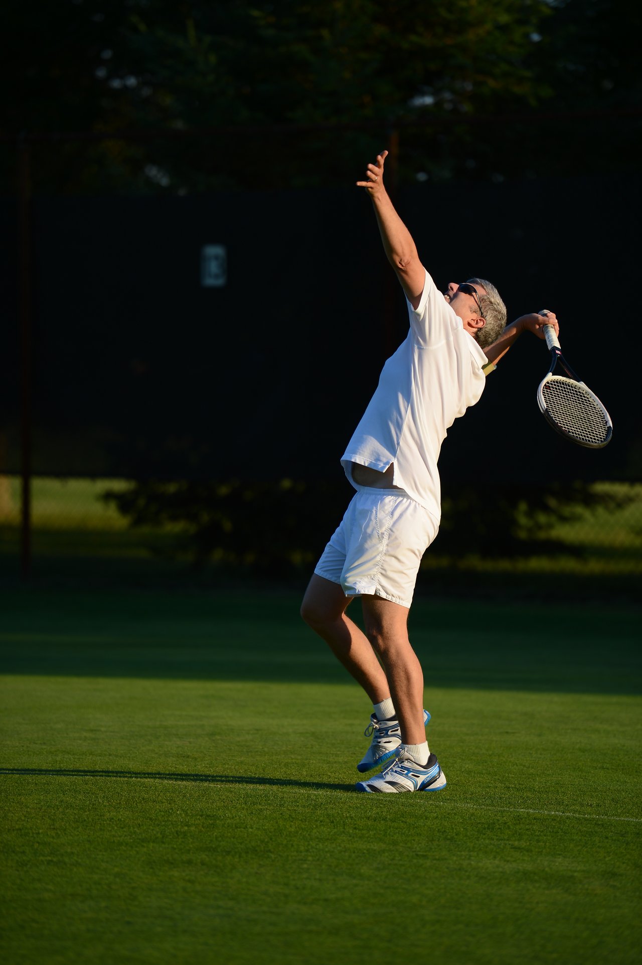 A tennis player in white clothing prepares to serve, tossing the ball and swinging the racket on a grass court.