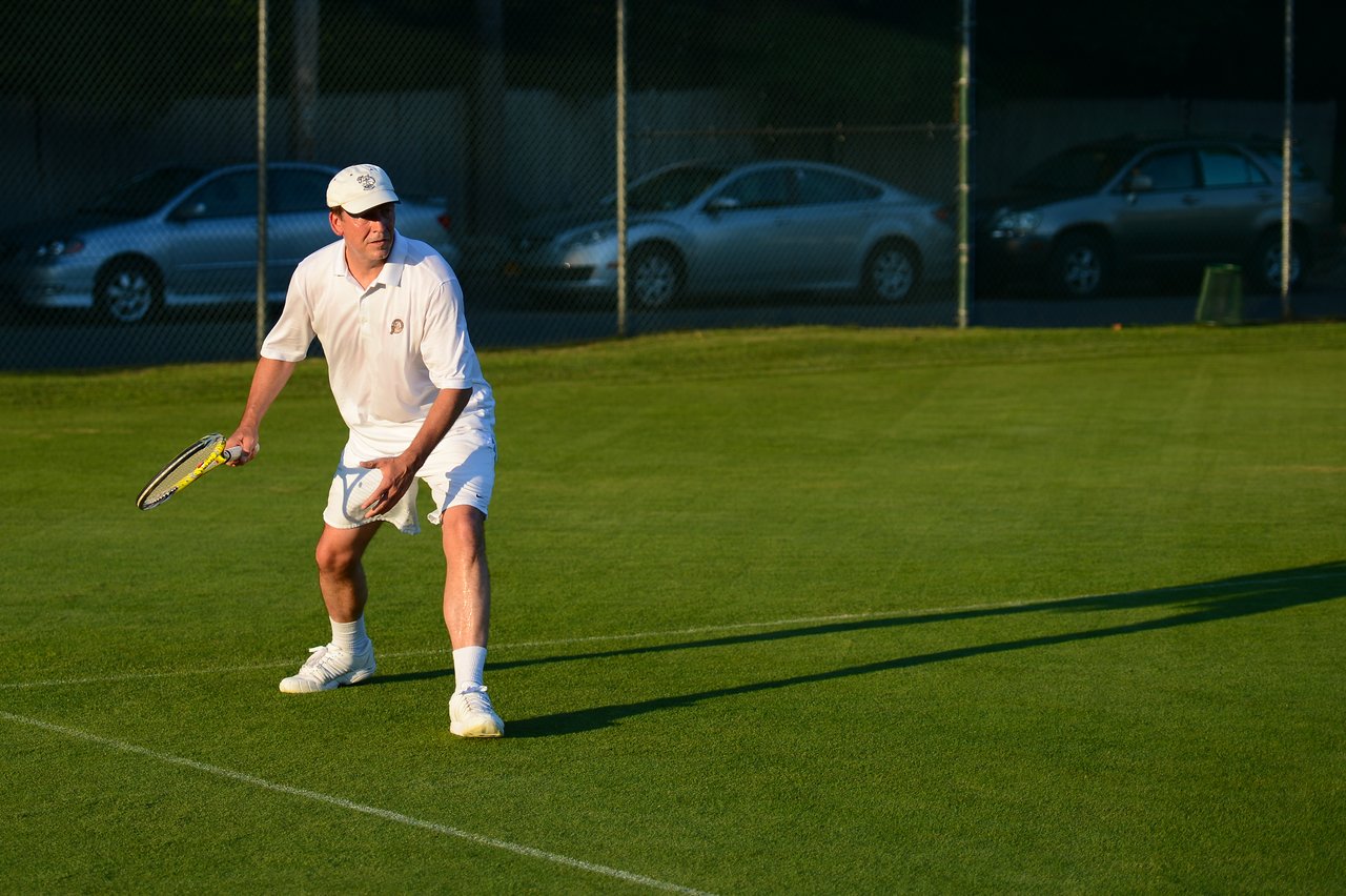 A tennis player in white attire prepares to receive a serve on a grass court, holding a racket.