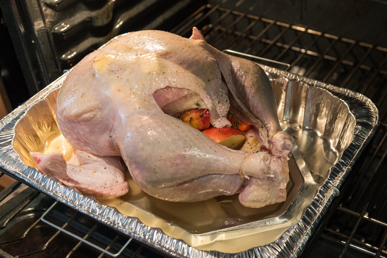 A raw, stuffed turkey in a roasting pan is placed inside an oven for cooking.