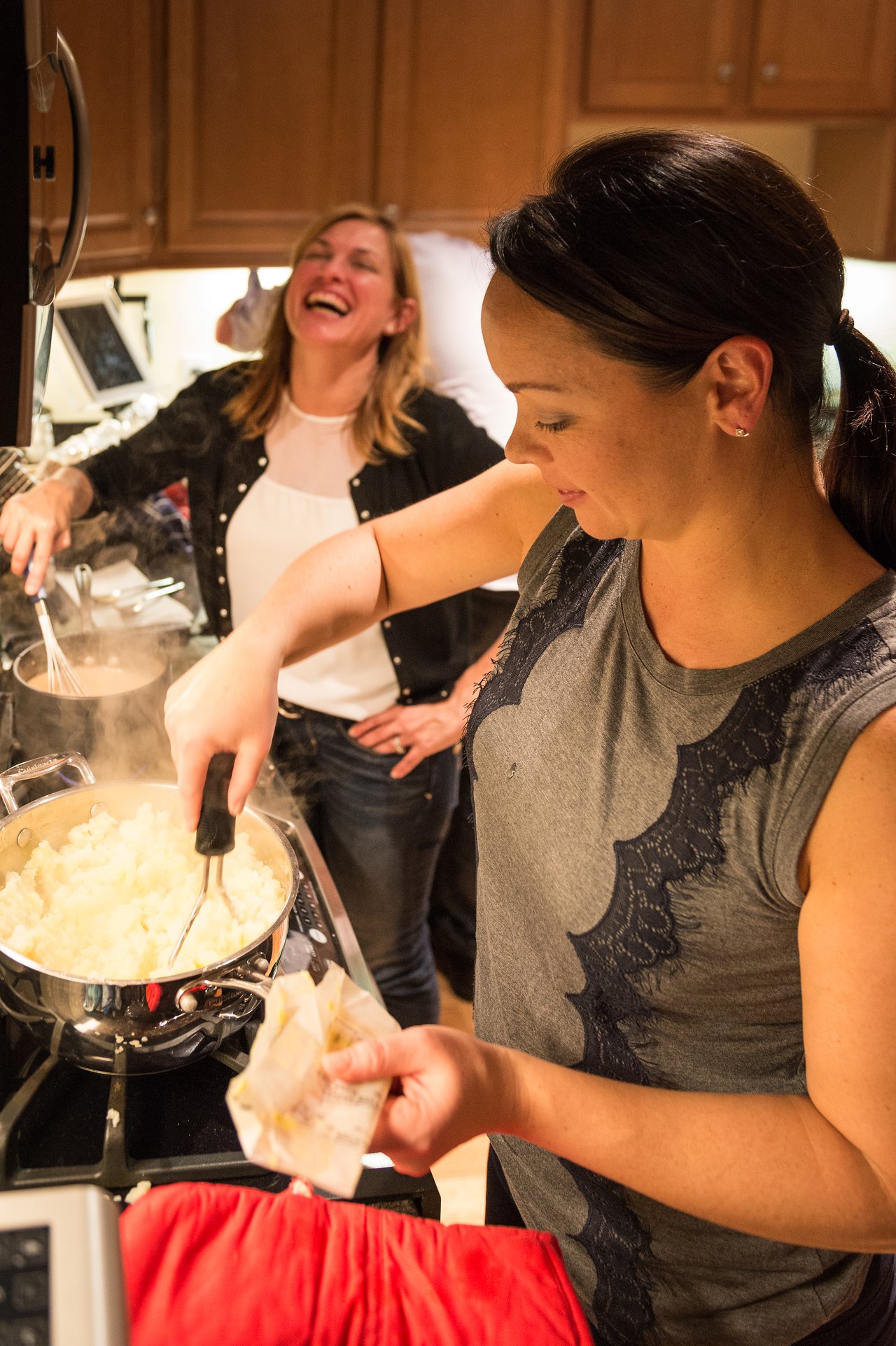 A woman mashes potatoes on the stove while another woman in the background laughs and talks.