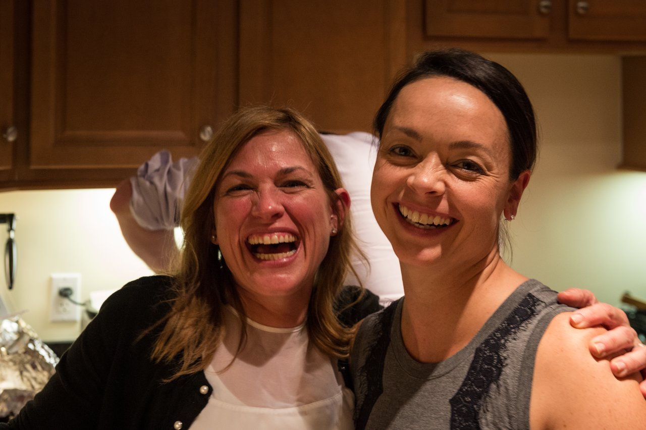 Two smiling women pose together in a kitchen, one with her arm around the other.