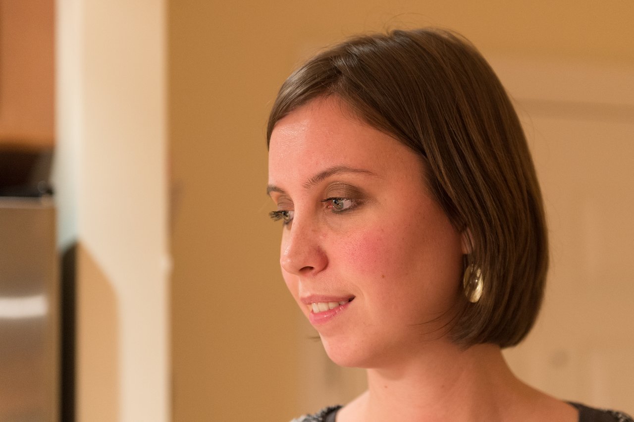 A woman with short brown hair and earrings looks to the side in a warmly lit indoor setting.
