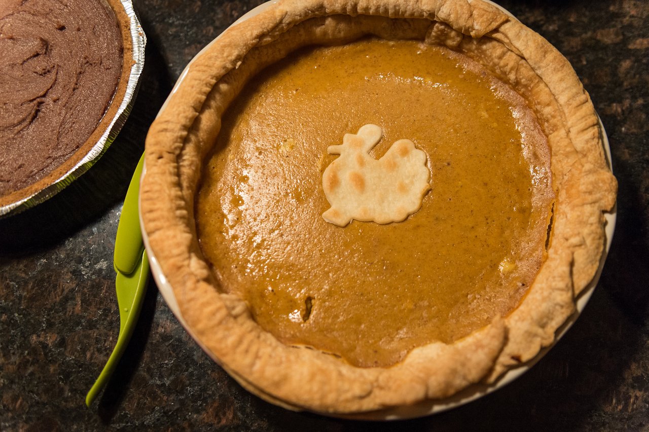 A pumpkin pie with a turkey-shaped crust decoration sits on a counter, next to a chocolate pie.