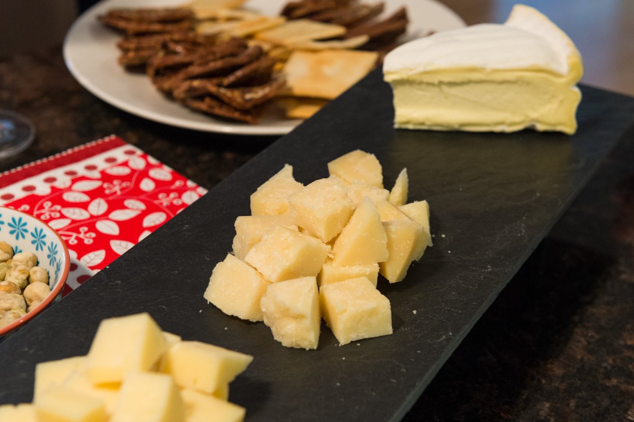 A cheese platter with cubed and sliced cheeses, crackers, and a bowl of snacks on a dark serving board.