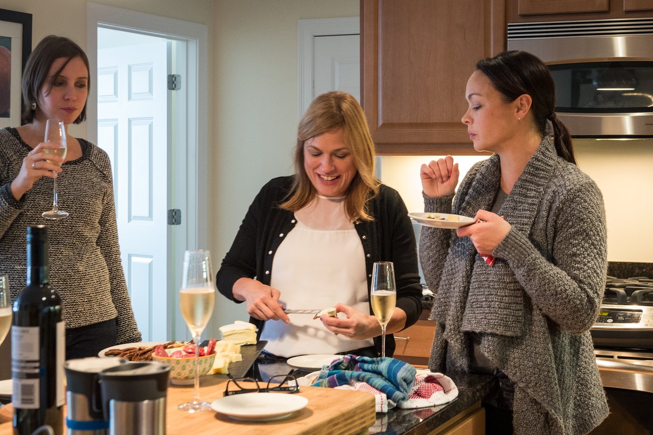 Three women in a kitchen enjoying food and drinks, with one slicing cheese and others eating and drinking.