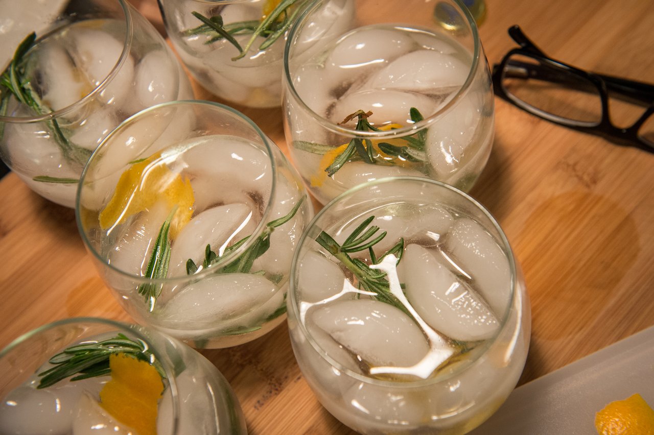 Several glasses filled with ice, rosemary, and citrus peel sit on a wooden surface, with glasses nearby.