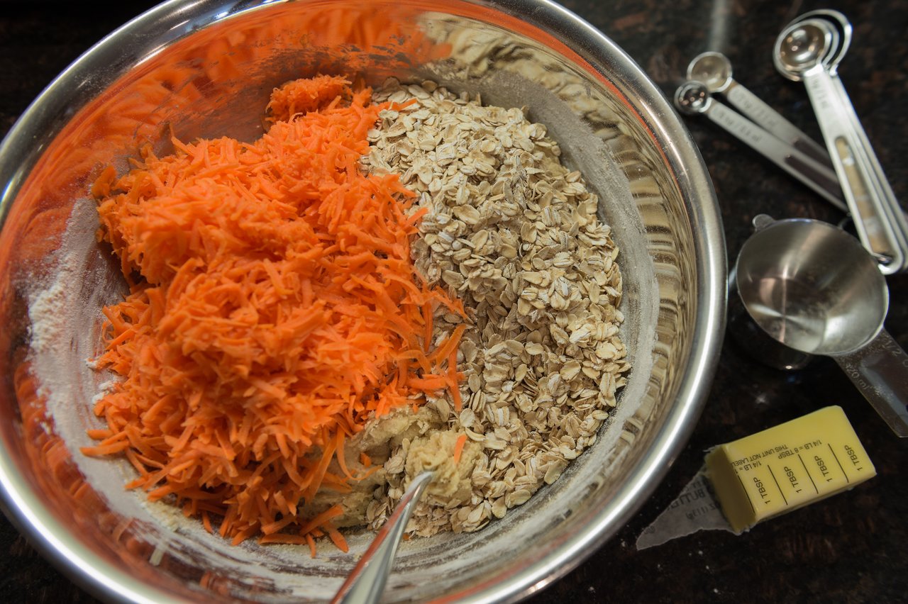 A mixing bowl with shredded carrots, oats, and flour, with measuring tools and butter nearby for baking carrot cake cookies.