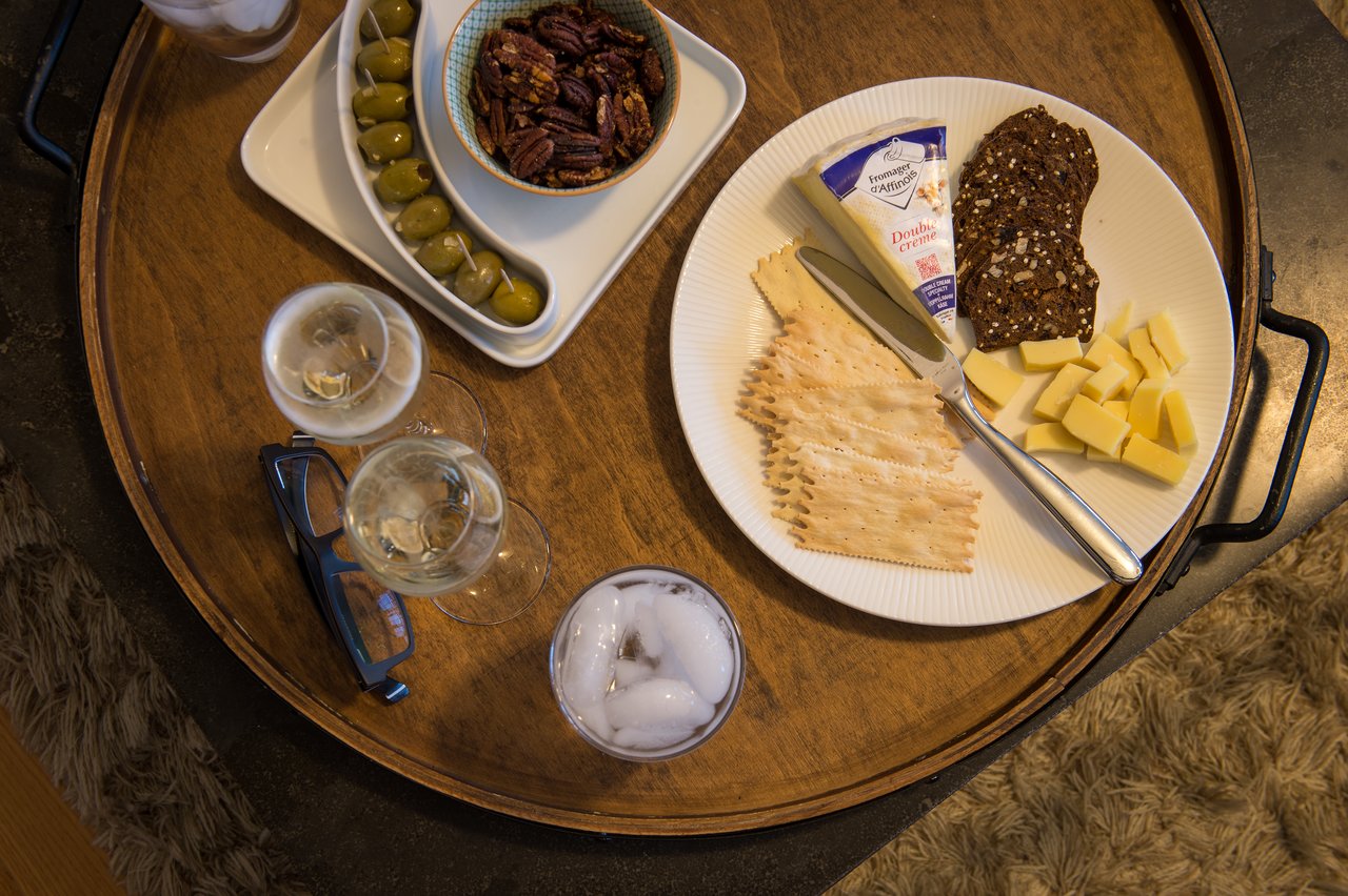 A wooden tray with cheese, crackers, nuts, olives, and drinks, set for a gathering.