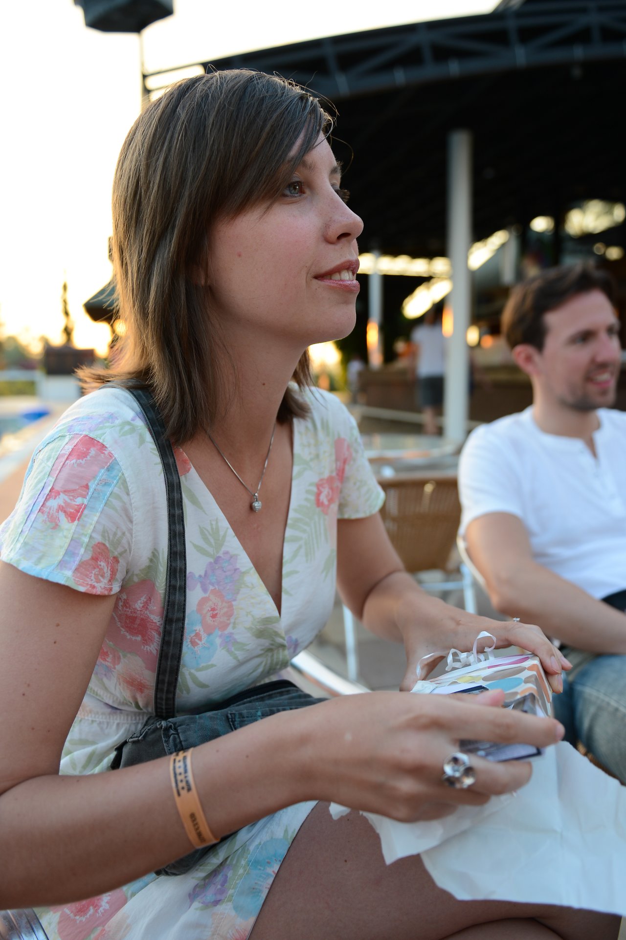 A woman in a floral dress holds a wrapped gift while sitting outdoors, with a man seated behind her.