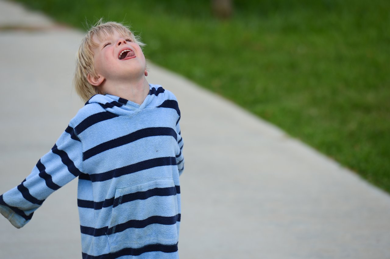 A child in a striped hoodie tilts their head back and smiles while standing on a paved path.