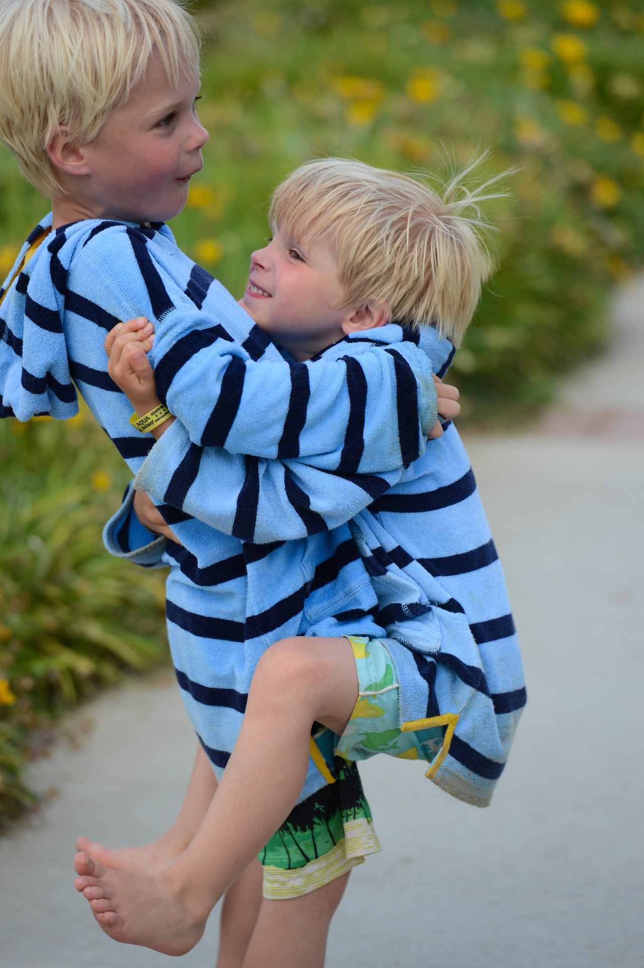 An older child in a striped hoodie lifts a younger child wearing matching clothes on a paved path.